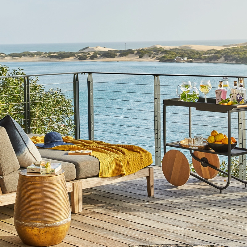 Perfect Hideaways: A stylish wooden deck with cushioned loungers, a golden side table, drinks trolley, and lanterns overlooks the sparkling estuary and sand dunes beyond, inviting relaxed outdoor living. St Austel, Kenton-on-Sea