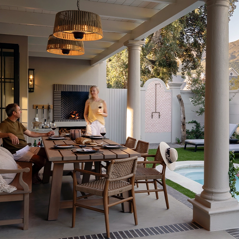 Perfect Hideaways: Covered veranda for outdoor dining with a long timber table, woven chairs and bench beneath woven pendants, a built‑in braai glowing, and a slim plunge pool edged with loungers, roses and a pink‑tiled outdoor shower, with mountain views and two guests enjoying a glass of wine, Franschhoek, Pink Door