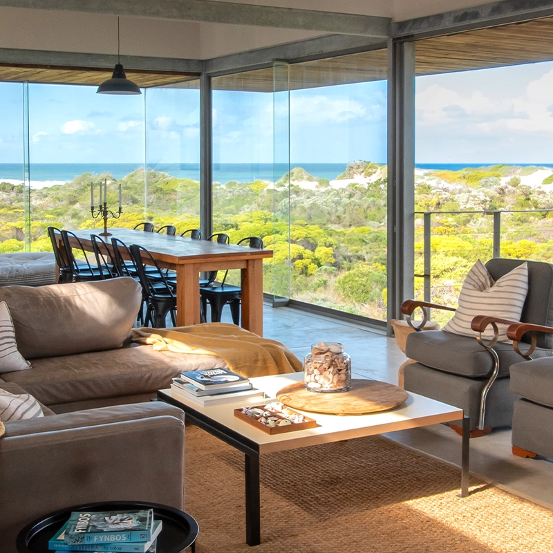 Perfect Hideaways: Open-plan lounge and dining area with floor-to-ceiling glass framing fynbos dunes and ocean views; leather corner sofa with mustard throw, two grey armchairs with striped cushions, a long wooden dining table with black chairs, pendant lights and a coffee table with books and shells, Betty's Bay, Westdunes