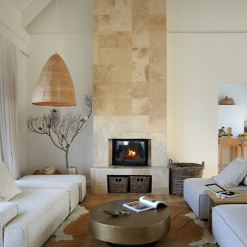 Perfect Hideaways: serene open-plan lounge with cream bouclé sofas facing a stone-clad fireplace, rattan pendant and woven baskets, round brass coffee table on a hide rug, and a light-wood kitchen beyond with sea views through sliding doors, Blanc de Blanc, Yzerfontein