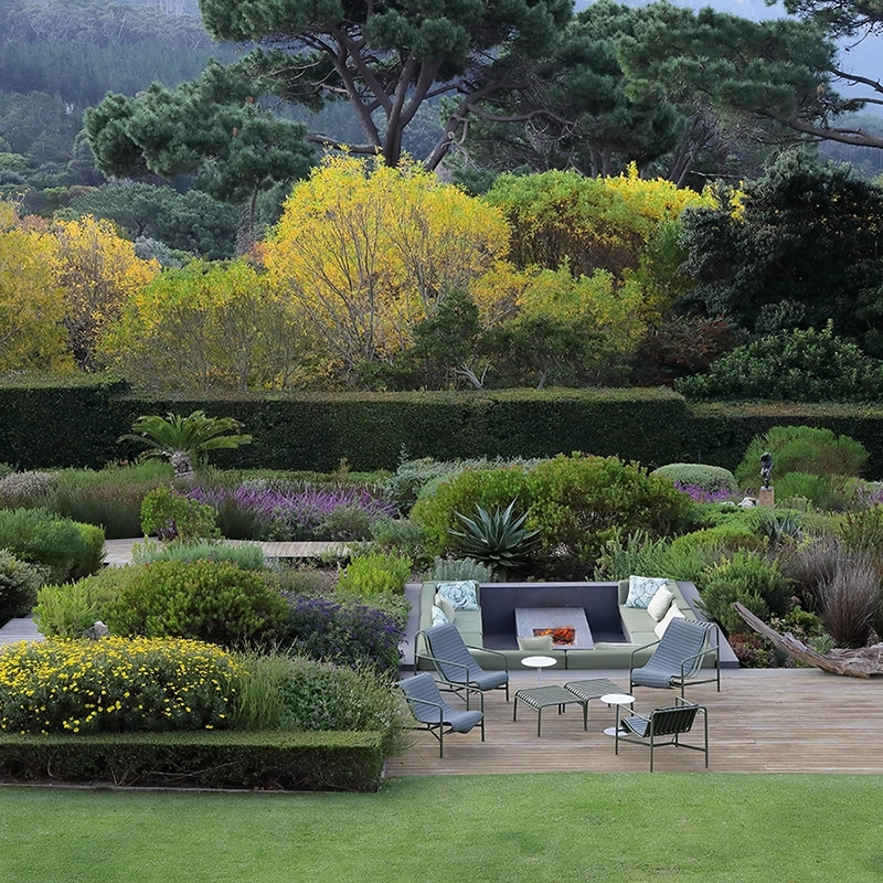 Perfect Hideaways: A beautifully landscaped garden with lush greenery, vibrant wildflowers, and manicured hedges surrounds an inviting outdoor seating area with a firepit. The scene is framed by towering trees and distant mountain views, creating a tranquil retreat. Dawn Villa, Constantia.