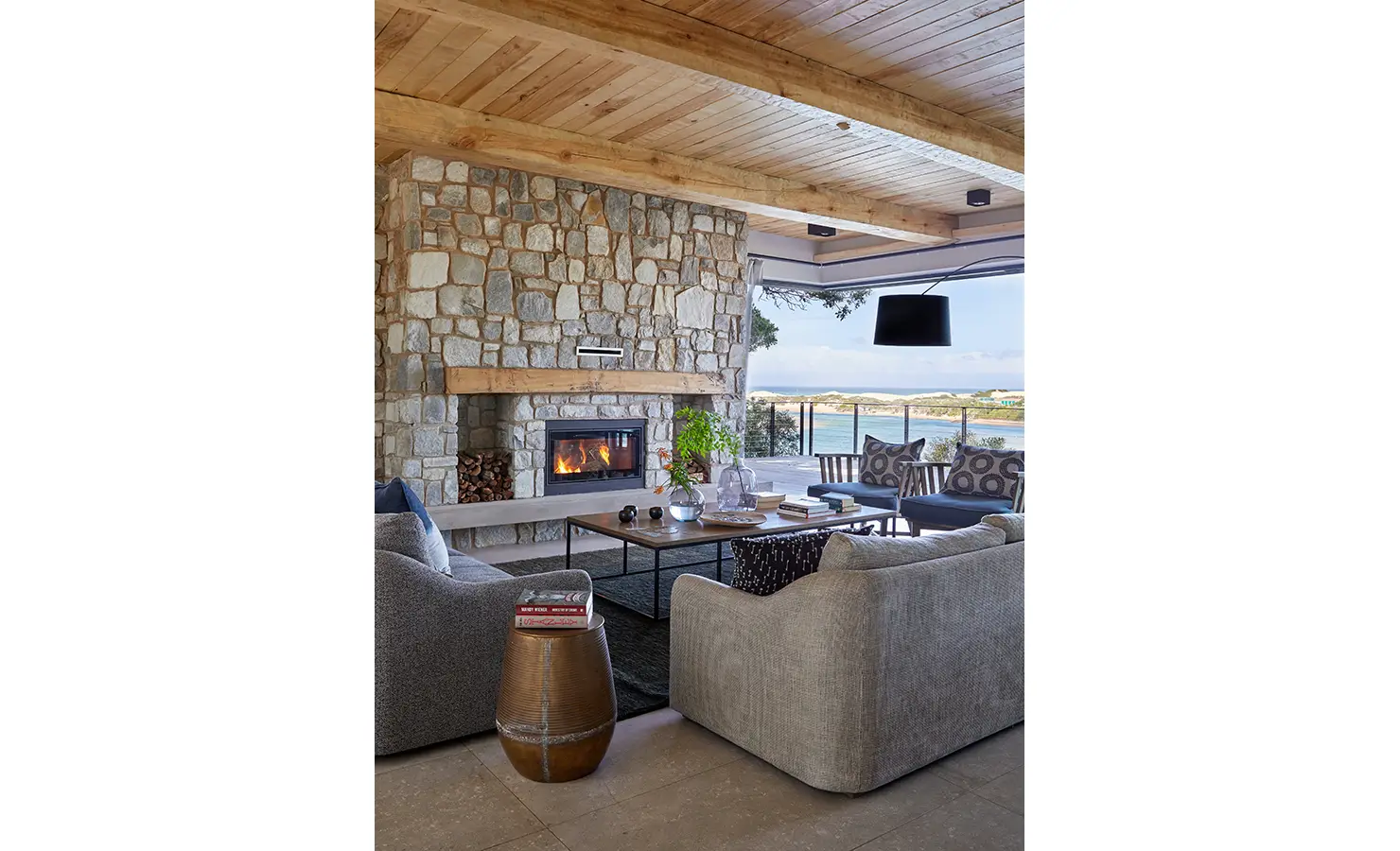 Perfect Hideaways: A spacious indoor lounge with soft grey sofas, a roaring stone fireplace, and panoramic views across the estuary through glass doors. St Austel, Kenton-on-Sea