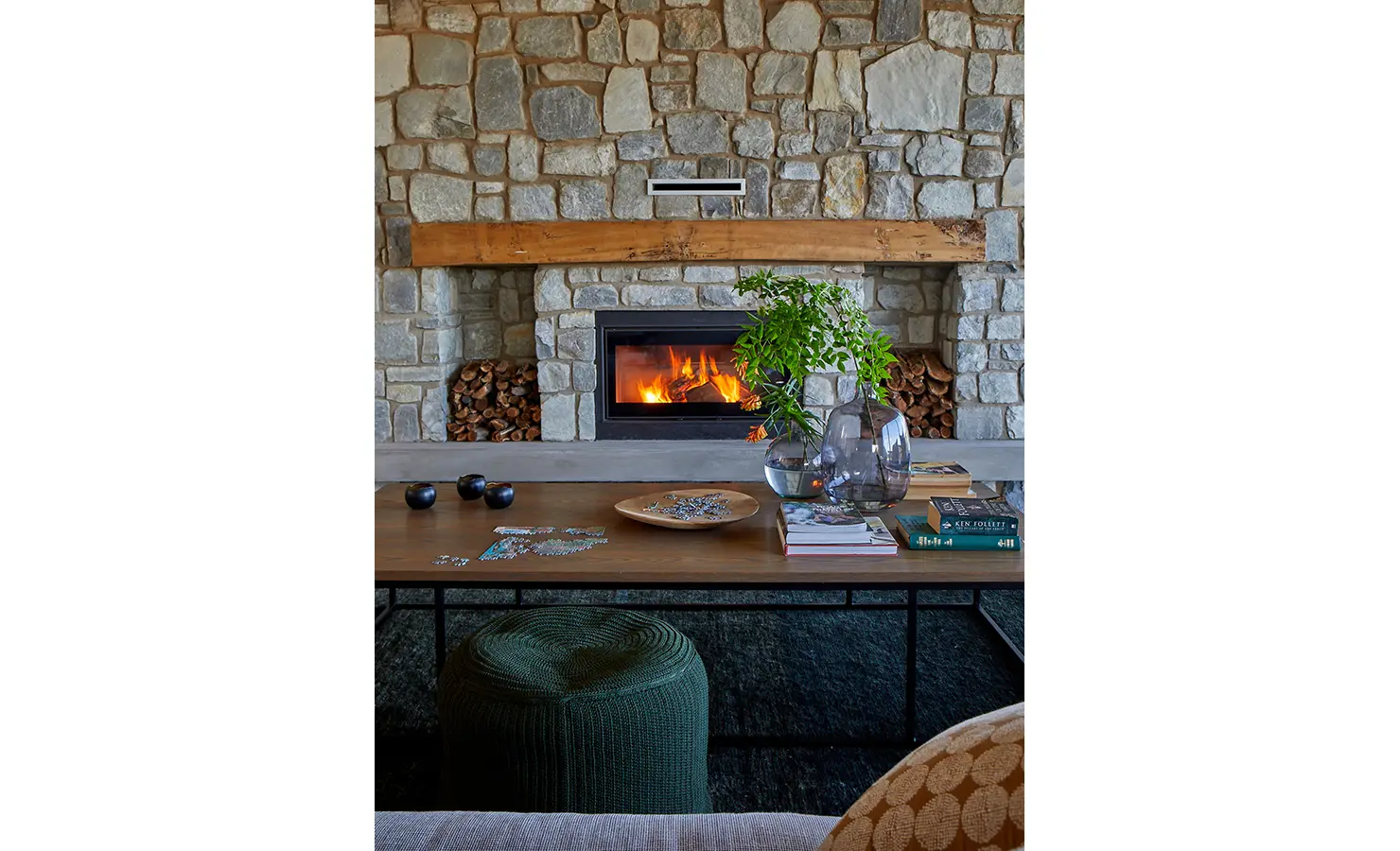 Perfect Hideaways: A close-up of a modern stone fireplace with a roaring fire, flanked by stacked logs, and a wooden coffee table styled with books, puzzles, and vases. St Austel, Kenton-on-Sea