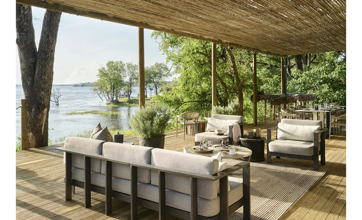 Perfect Hideaways: Shaded riverside lounge under a reed-roof pergola with cushioned sofas and dining area opening to uninterrupted Zambezi views. Hippo Creek Villa, Zambezi National Park.