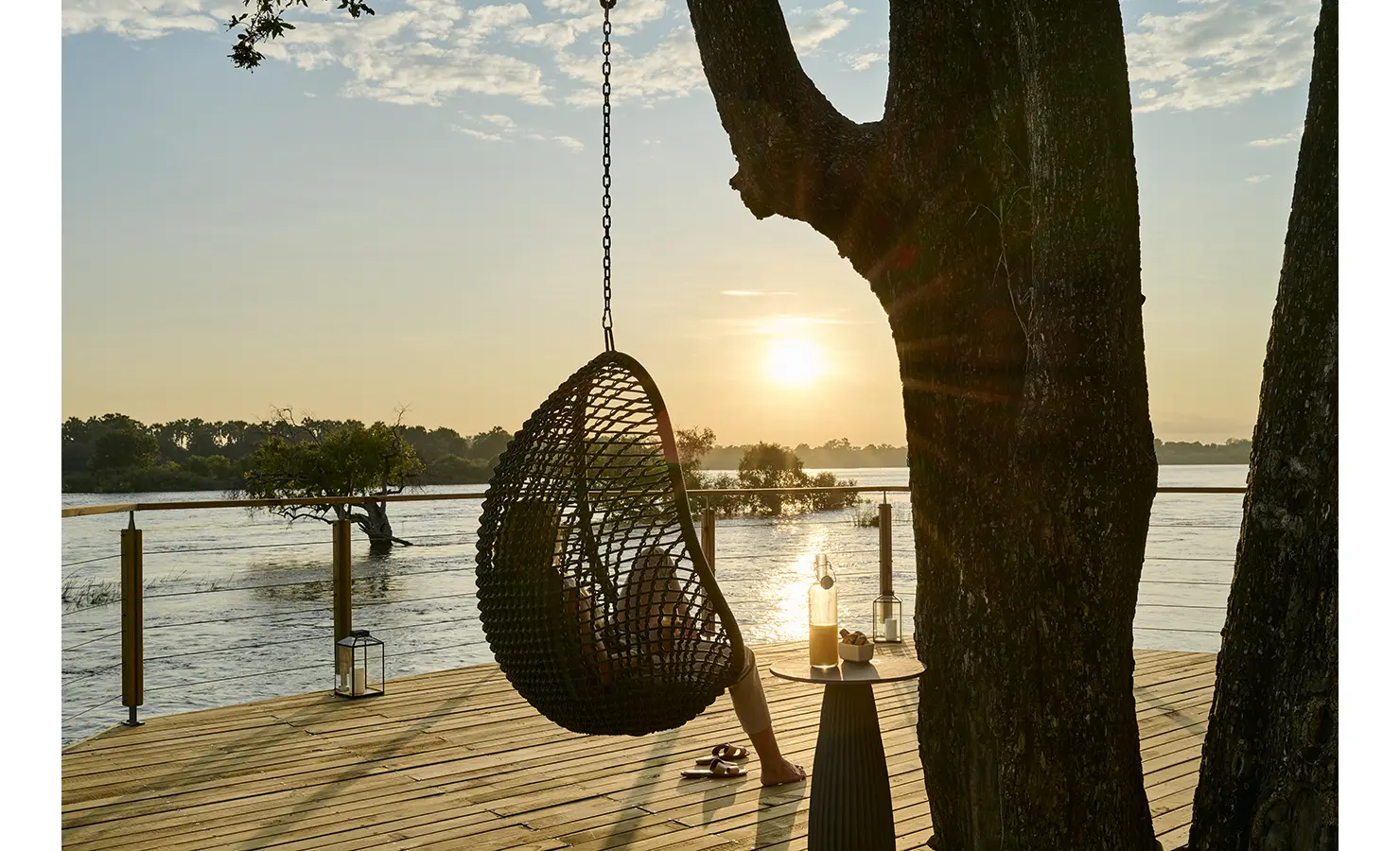 Perfect Hideaways: Hanging basket chair beneath a riverside tree at sunset with lantern and drinks on the deck, Rapid's View Villa, Zambezi National Park