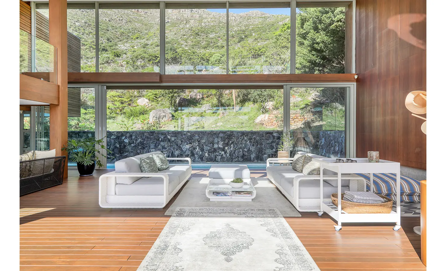 Perfect Hideaways: Double‑height living room with white sofas facing floor‑to‑ceiling glass to the mountain slope and a lap pool beyond a stone wall, Spa House, Hout Bay