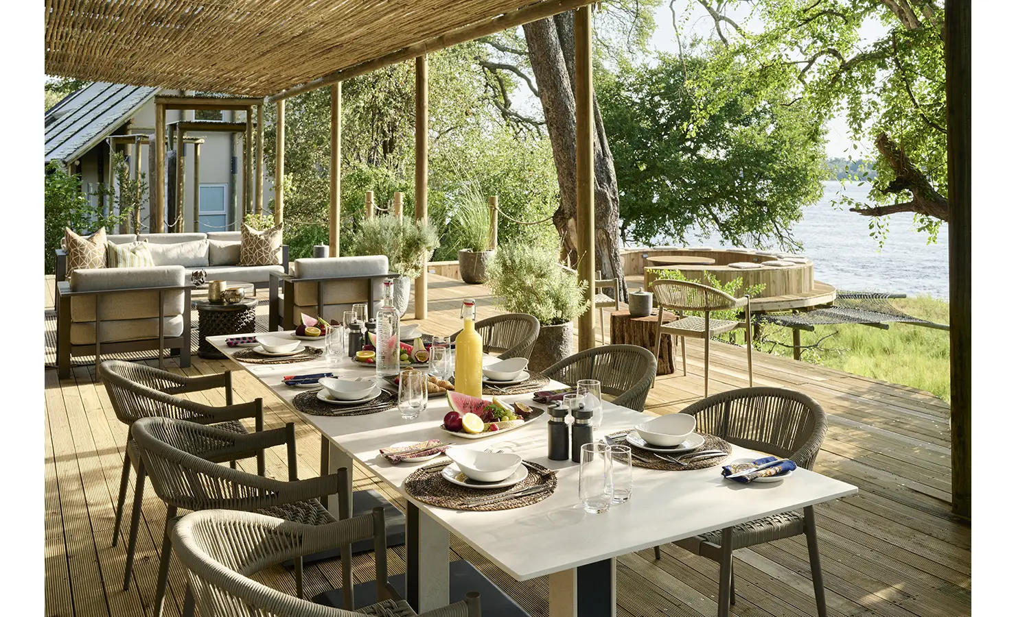 Perfect Hideaways: Al fresco dining deck laid for lunch beneath a reed canopy, with cushioned lounge and circular timber hot tub overlooking the Zambezi. Hippo Creek Villa, Zambezi National Park.