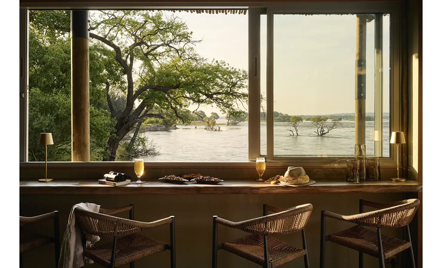 Perfect Hideaways: Indoor window bar with live-edge counter and woven chairs gazing onto a tree-lined river at golden hour, Rapid's View Villa, Zambezi National Park