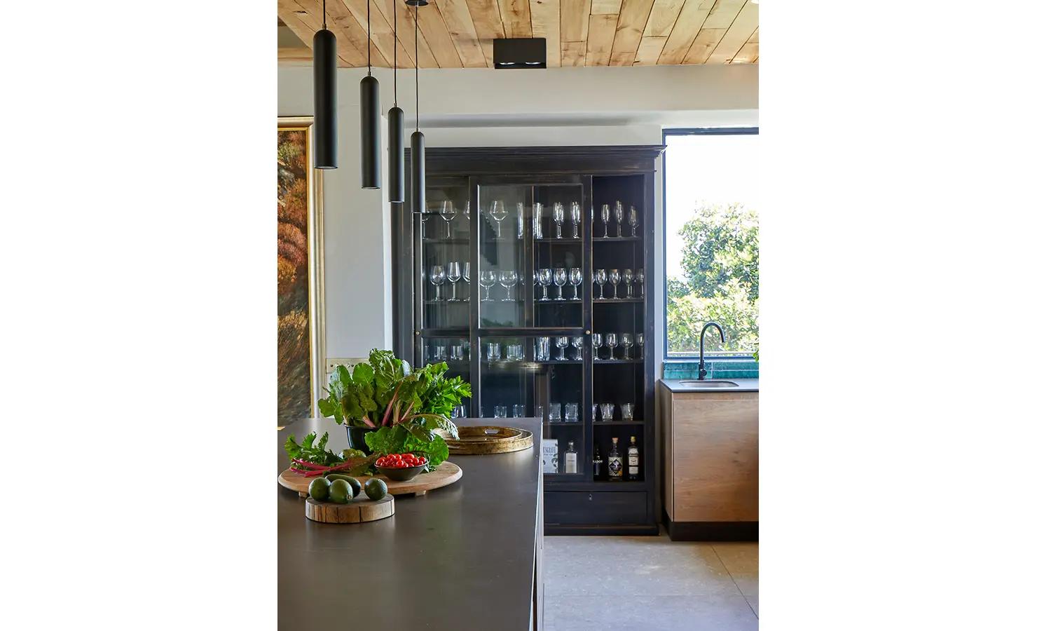 Perfect Hideaways: A black-framed glass cabinet filled with crystalware stands beside a sunlit kitchen island topped with fresh vegetables and elegant trays. St Austel, Kenton-on-Sea