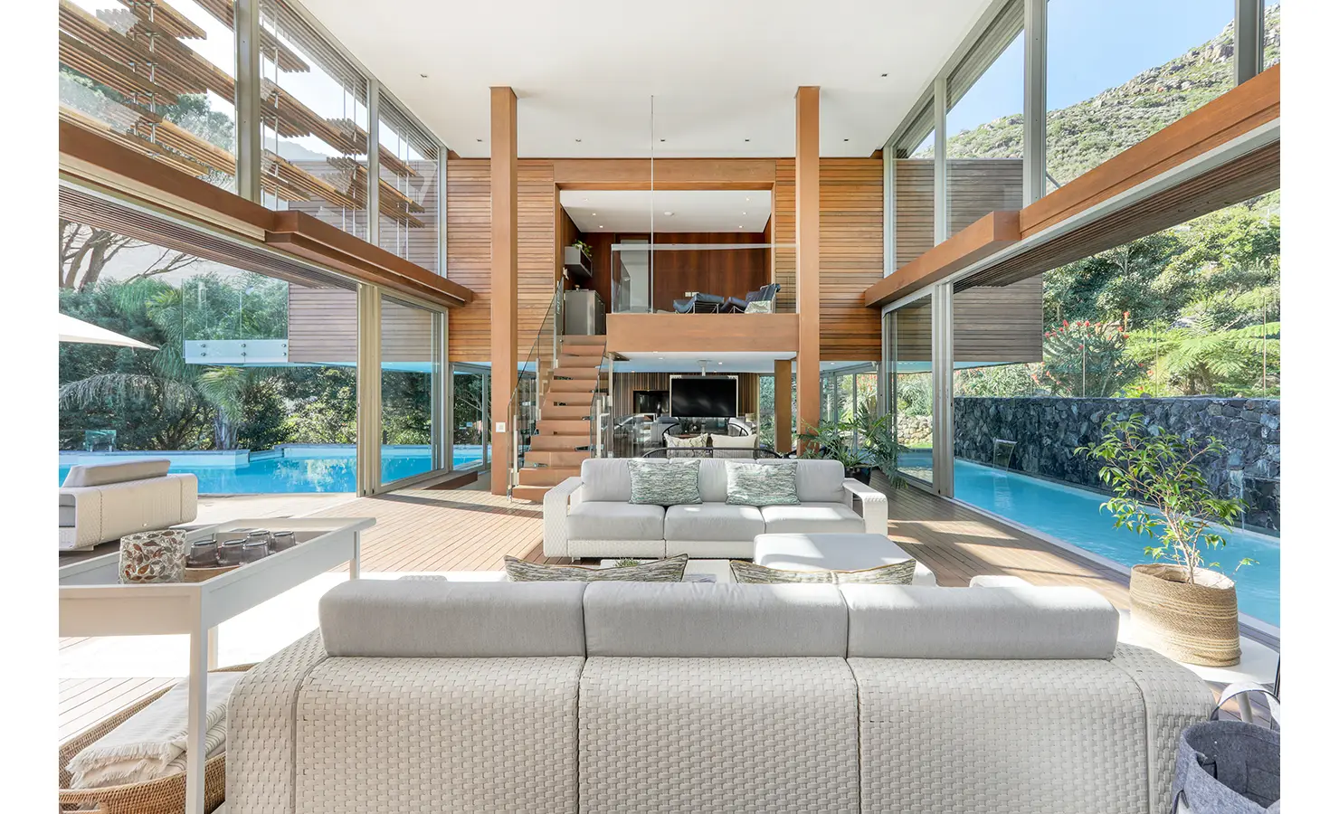 Perfect Hideaways: Central double‑volume space with timber columns and stair to a mezzanine, glass walls opening to a wraparound pool and decks, Spa House, Hout Bay