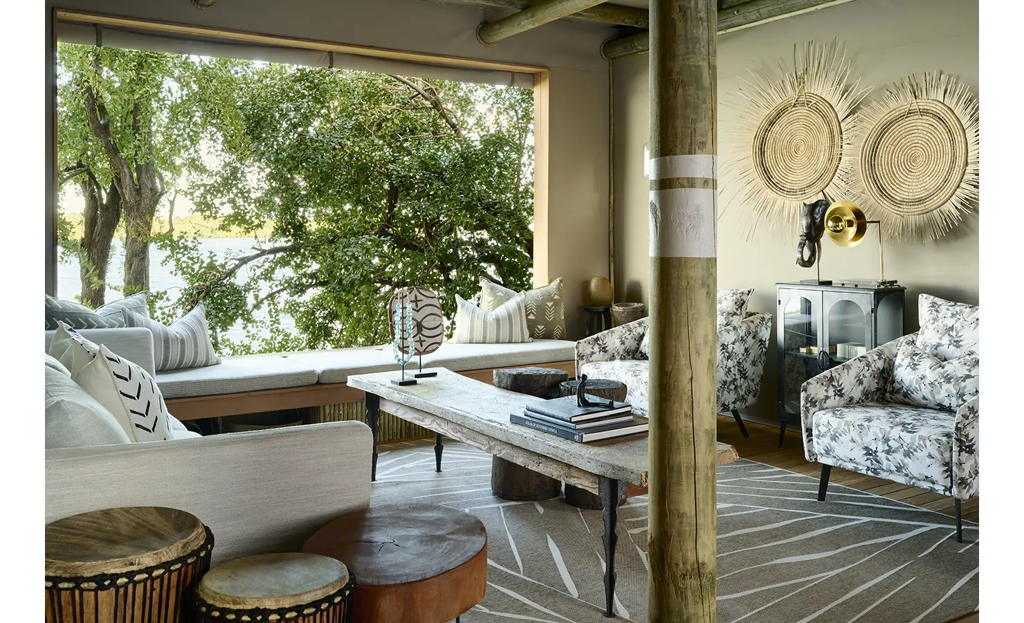 Perfect Hideaways: Cosy lounge nook with window seat, patterned armchairs and woven wall art framing leafy river views. Hippo Creek Villa, Zambezi National Park.