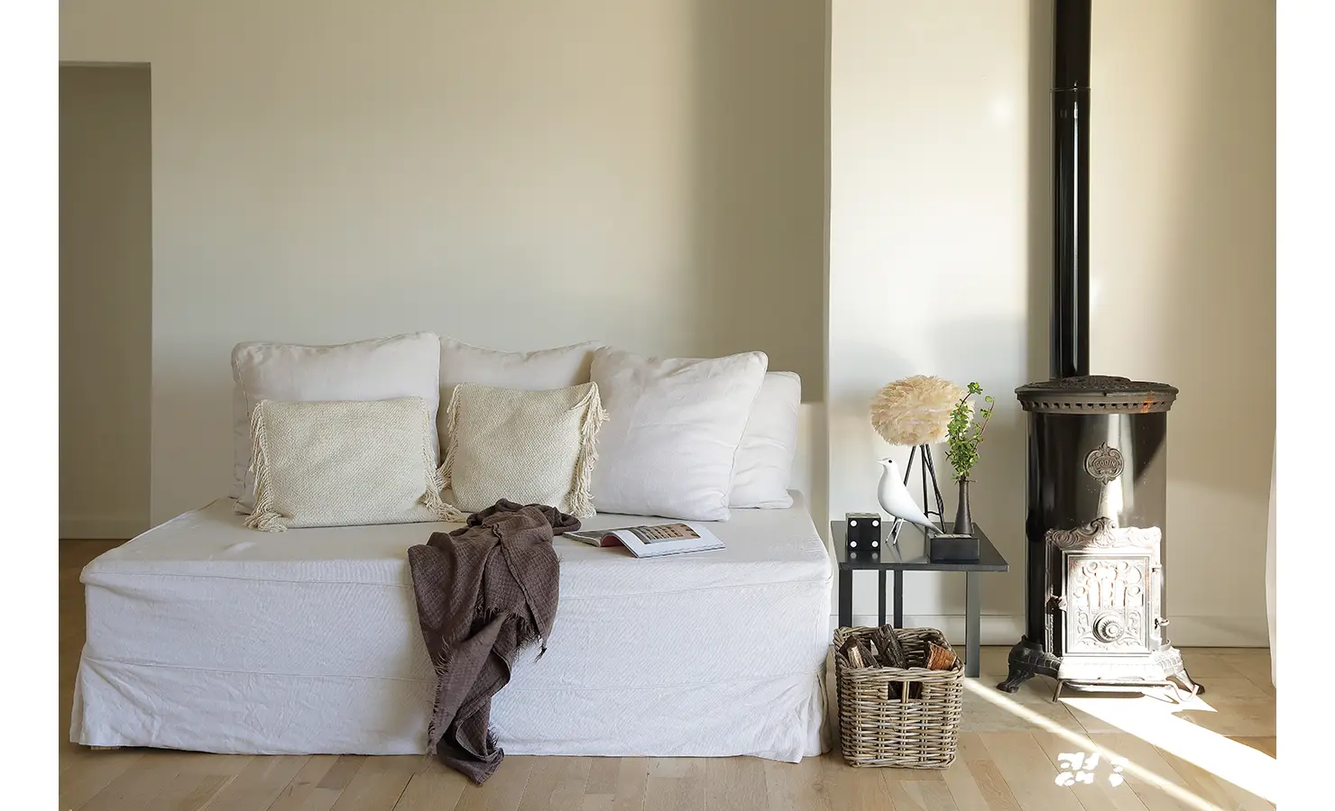 Perfect Hideaways: tranquil snug with a white daybed, textured cushions and an antique black wood-burning stove, wicker log basket and simple side table, Blanc de Blanc, Yzerfontein