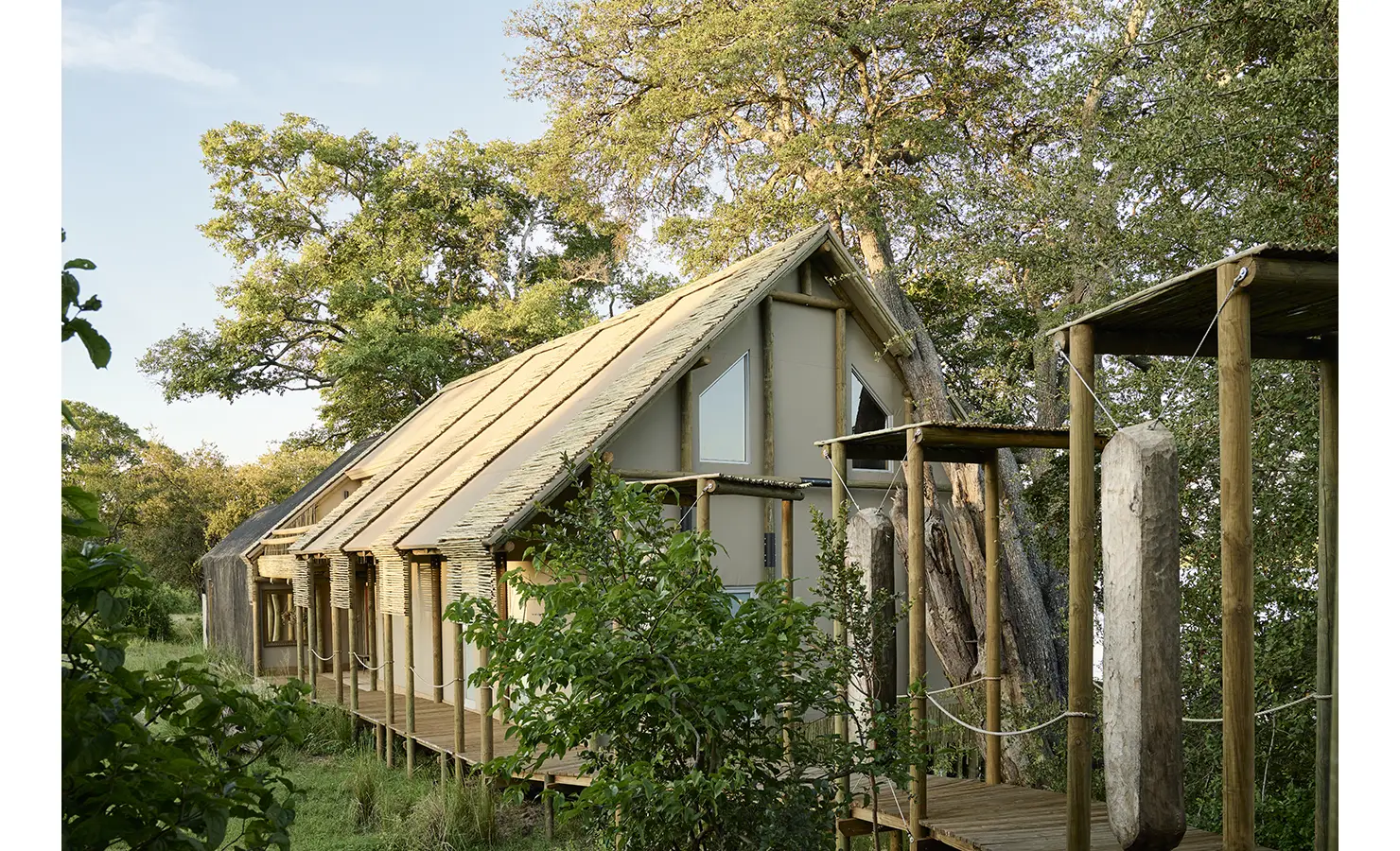 Perfect Hideaways: Exterior of the thatched, timber-framed villa on raised boardwalks, blending into riverine woodland at golden hour. Hippo Creek Villa, Zambezi National Park.