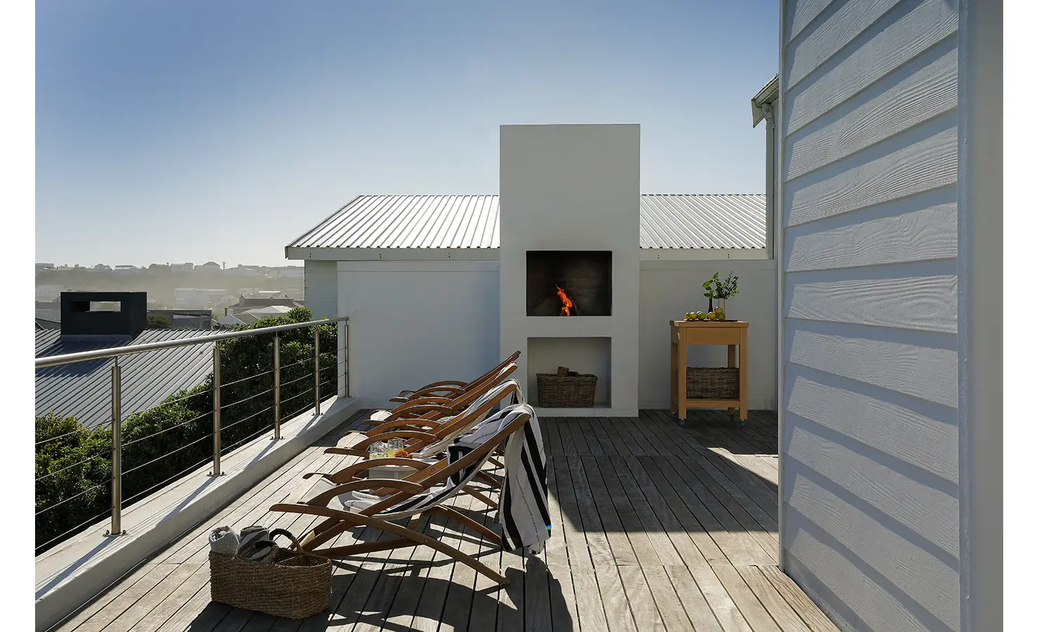 Perfect Hideaways: upper deck with a row of wooden sling loungers facing a built-in outdoor fireplace, stainless-steel cable balustrade and coastal rooftops under a clear sky, Blanc de Blanc, Yzerfontein