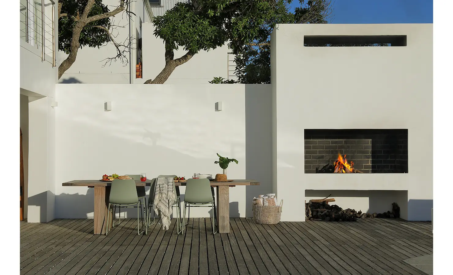 Perfect Hideaways: sheltered courtyard deck with rustic dining table and sage chairs beside a modern white outdoor fireplace, logs stacked ready for the braai, Blanc de Blanc, Yzerfontein