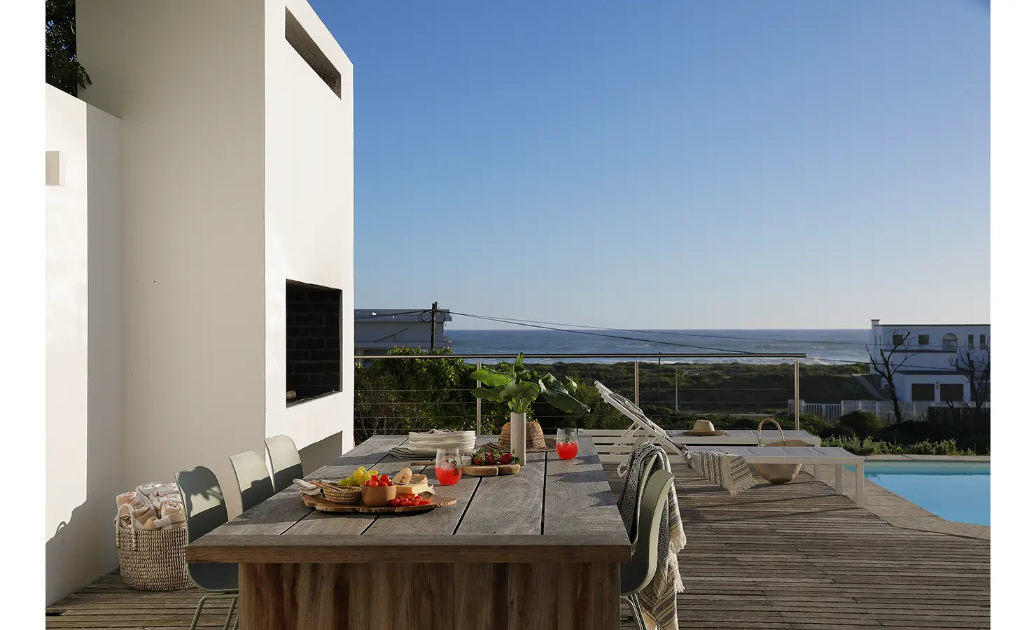Perfect Hideaways: alfresco dining table dressed with fruit and drinks beside the built-in fireplace, pool terrace and ocean beyond the glass balustrade, Blanc de Blanc, Yzerfontein