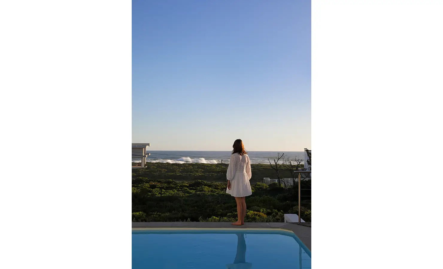 Perfect Hideaways: guest standing at the pool’s edge gazing over coastal fynbos to breaking waves beneath a cloudless evening sky, Blanc de Blanc, Yzerfontein