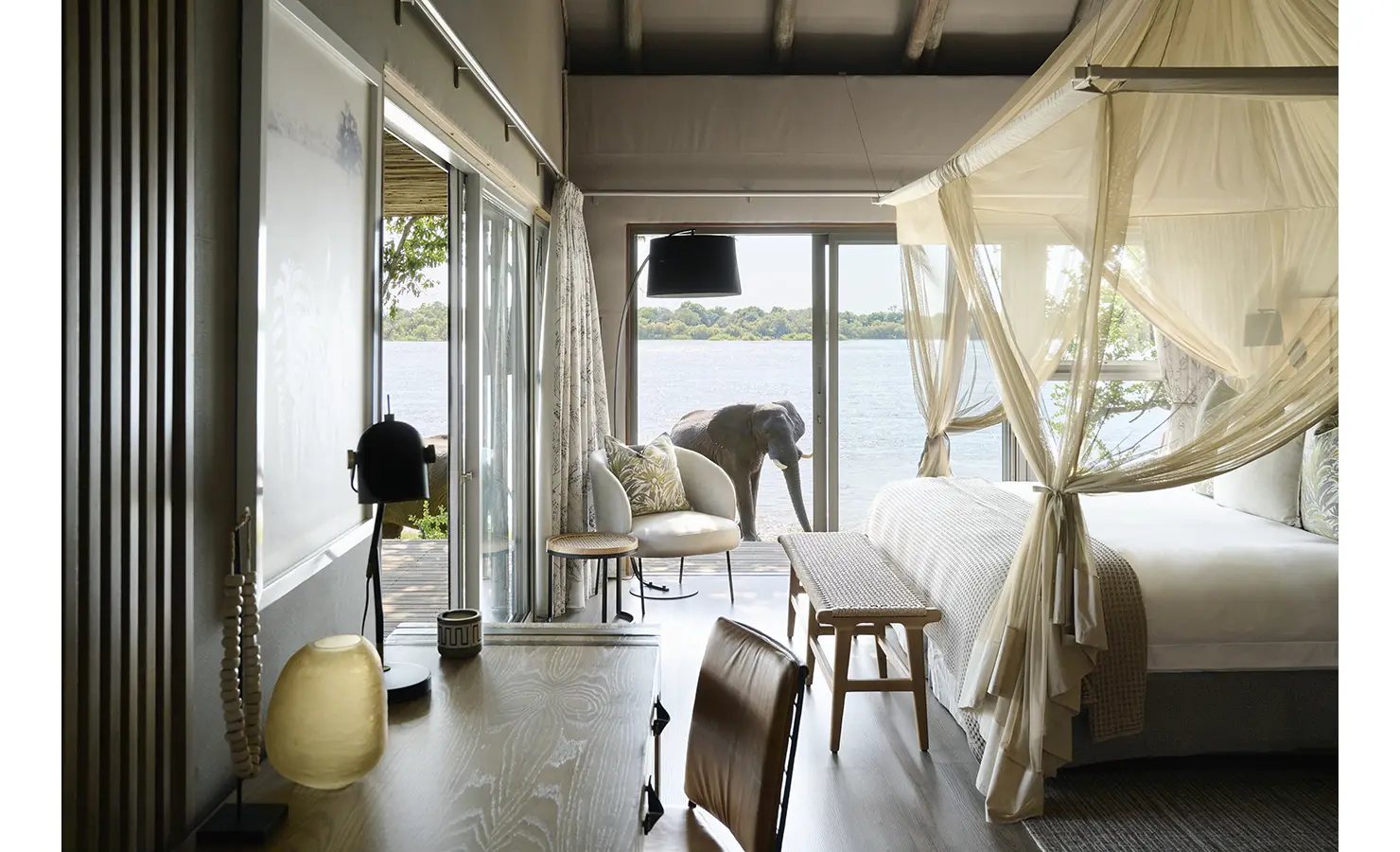Perfect Hideaways: Light-filled suite with canopy bed and sliding doors opening onto the deck, with an elephant grazing at the river’s edge. Hippo Creek Villa, Zambezi National Park.