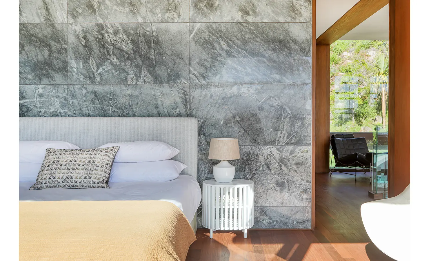 Perfect Hideaways: Bedroom detail with marble-clad feature wall, upholstered headboard, bedside lamp and view to a lounge through a timber portal, Spa House, Hout Bay