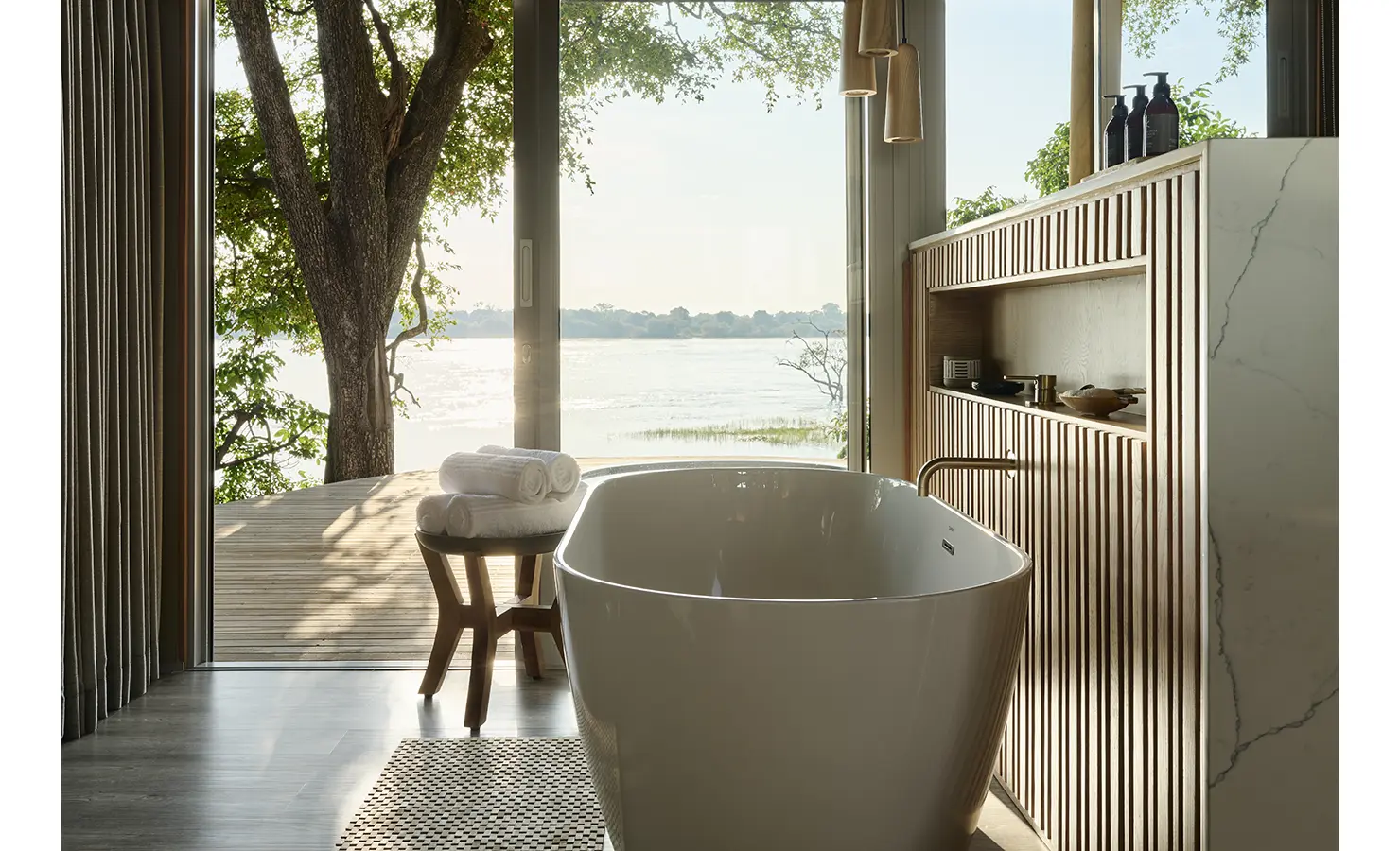 Perfect Hideaways: Indoor freestanding bathtub with timber feature shelving, large glass doors opening to the deck and sweeping Zambezi views. Hippo Creek Villa, Zambezi National Park.