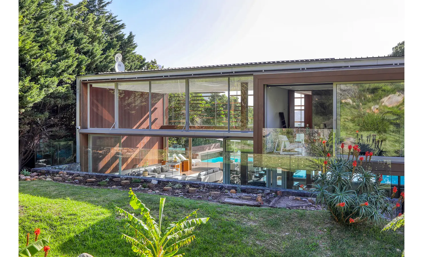 Perfect Hideaways: Garden elevation of the glass-and-timber pavilion showing double-height living room and upper suite, framed by aloes and lush greenery, Spa House, Hout Bay