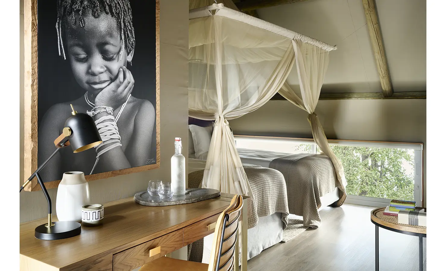 Perfect Hideaways: Cosy loft-style twin bedroom with canopy nets, writing desk and monochrome portrait artwork, large window with leafy river views. Hippo Creek Villa, Zambezi National Park.