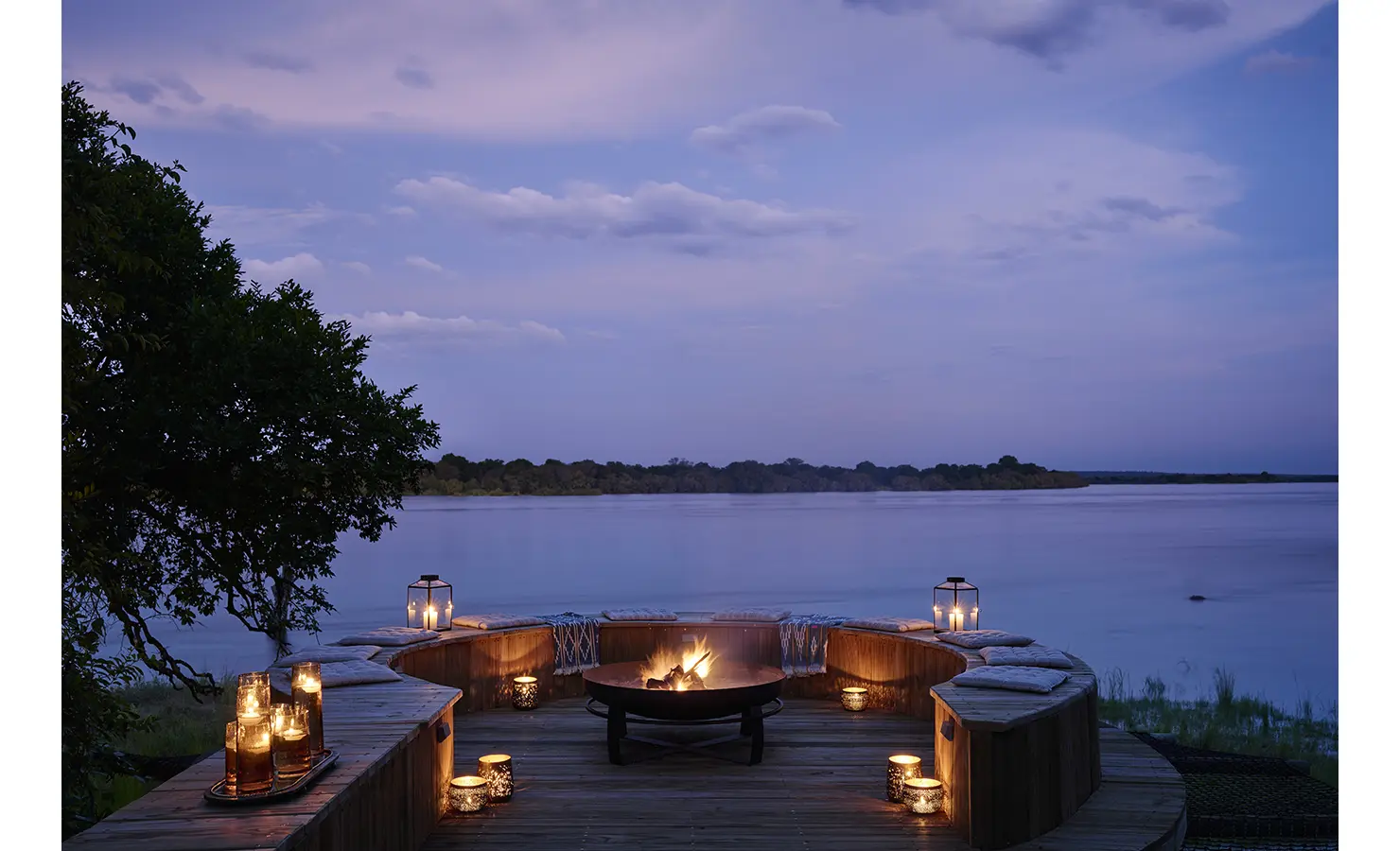 Perfect Hideaways: Curved riverside boma with timber seating and a central fire, lanterns aglow against the twilight Zambezi. Hippo Creek Villa, Zambezi National Park.