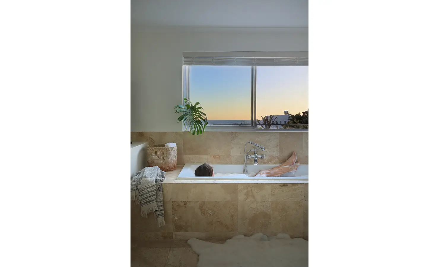 Perfect Hideaways: stone-tiled bathroom with built-in tub facing a sunset sea view, monstera leaf by the window, woven basket and soft rug, Blanc de Blanc, Yzerfontein