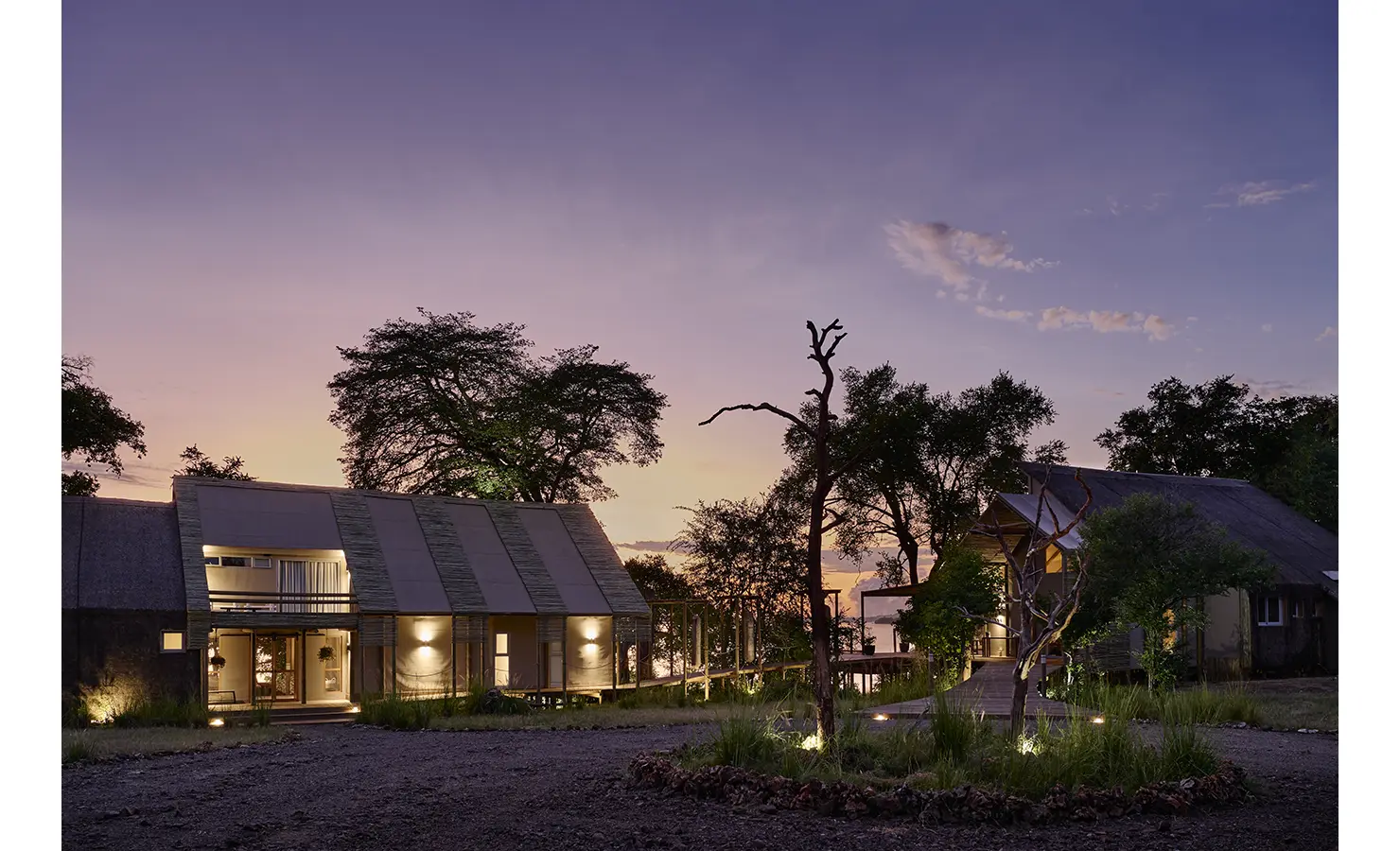 Perfect Hideaways: Dusk exterior of the thatched villa cluster, softly lit with boardwalks and trees framing glimpses of the river. Hippo Creek Villa, Zambezi National Park.