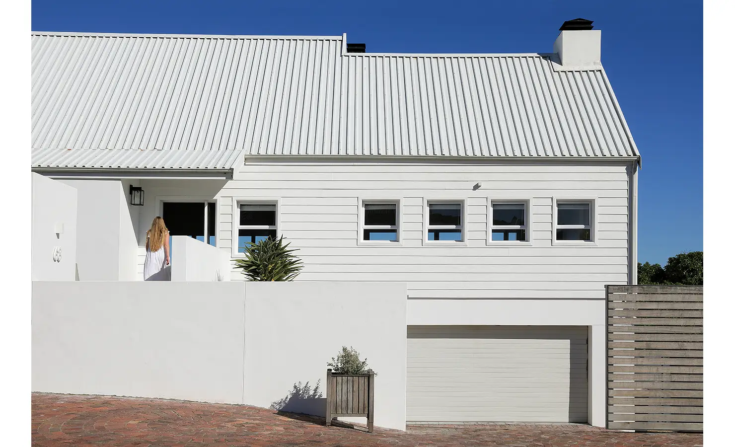Perfect Hideaways: coastal façade in white weatherboard with corrugated roof, row of square windows, garage below and a guest entering the front door, Blanc de Blanc, Yzerfontein