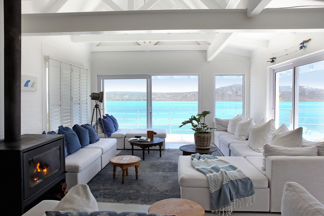 Perfect Hideaways: light-filled coastal living room with white sectional sofas, blue cushions and throws, a wood-burning stove and panoramic turquoise lagoon views framed by picture windows, Langebaan, Lagoon Lookout.
