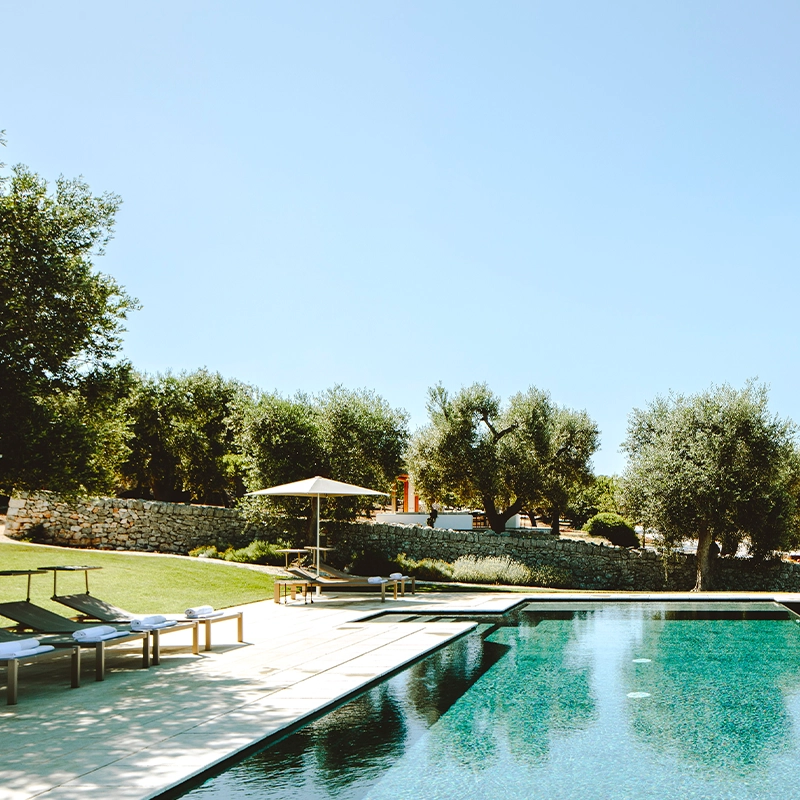 Perfect Hideaways: A serene view of a glistening turquoise swimming pool surrounded by olive trees, stone walls, and sun loungers with white rolled towels under large beige parasols, all set against a backdrop of trulli rooftops under a clear blue sky. Masseria Monopoli, Monopoli.