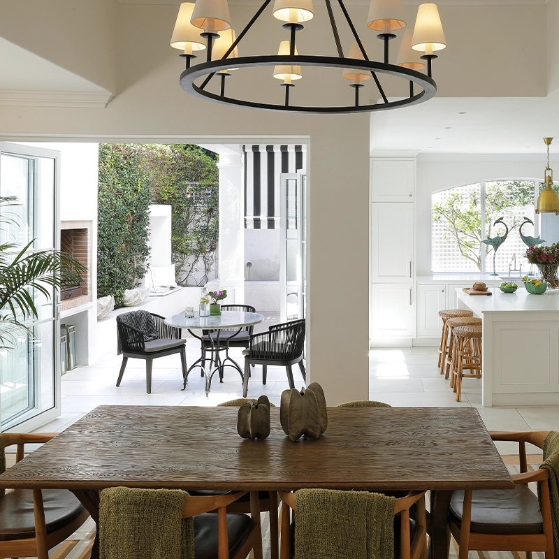 Perfect Hideaways: A warm and inviting open-plan dining, kitchen and patio space, where a rustic timber table is paired with modern chairs and a statement chandelier, leading out through folding glass doors to a bright courtyard with black woven seating and leafy vertical greenery. Fresnaye, The Townhouse.