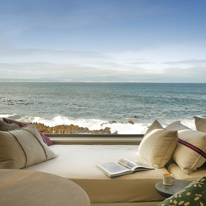 Perfect Hideaways: A serene ocean-facing daybed with soft linen cushions, a coffee cup, and an open book invites relaxation while waves crash against the rocks just beyond the glass window, offering uninterrupted sea views. The Keep, Hermanus.