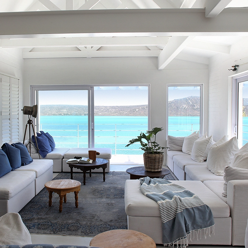 Perfect Hideaways: light-filled coastal living room with white sectional sofas, blue cushions and throws, a wood-burning stove and panoramic turquoise lagoon views framed by picture windows, Langebaan, Lagoon Lookout.