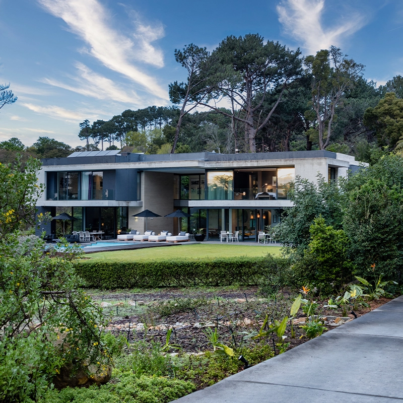 Perfect Hideaways: A striking contemporary villa with expansive glass walls, shaded terraces, and a manicured lawn leading to a pool, set against a lush forest backdrop in Constantia, House Nour.