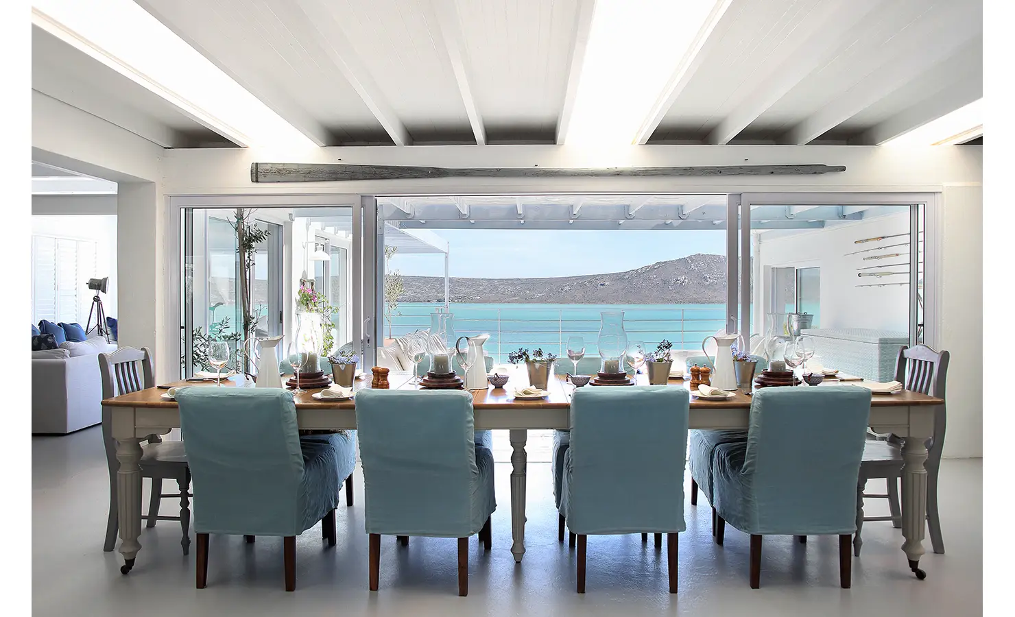 Perfect Hideaways: long dining table with blue slipcovered chairs set beside glass doors framing calm lagoon and mountain views, Langebaan, Lagoon Lookout.