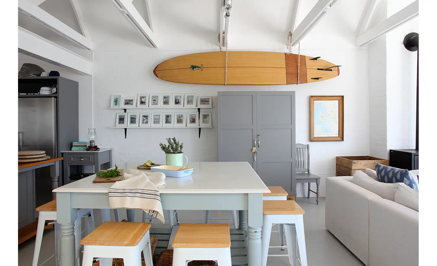 Perfect Hideaways: airy kitchen–living space with vintage longboard hung from beams, framed photos and a central island with stools, Langebaan, Lagoon Lookout.