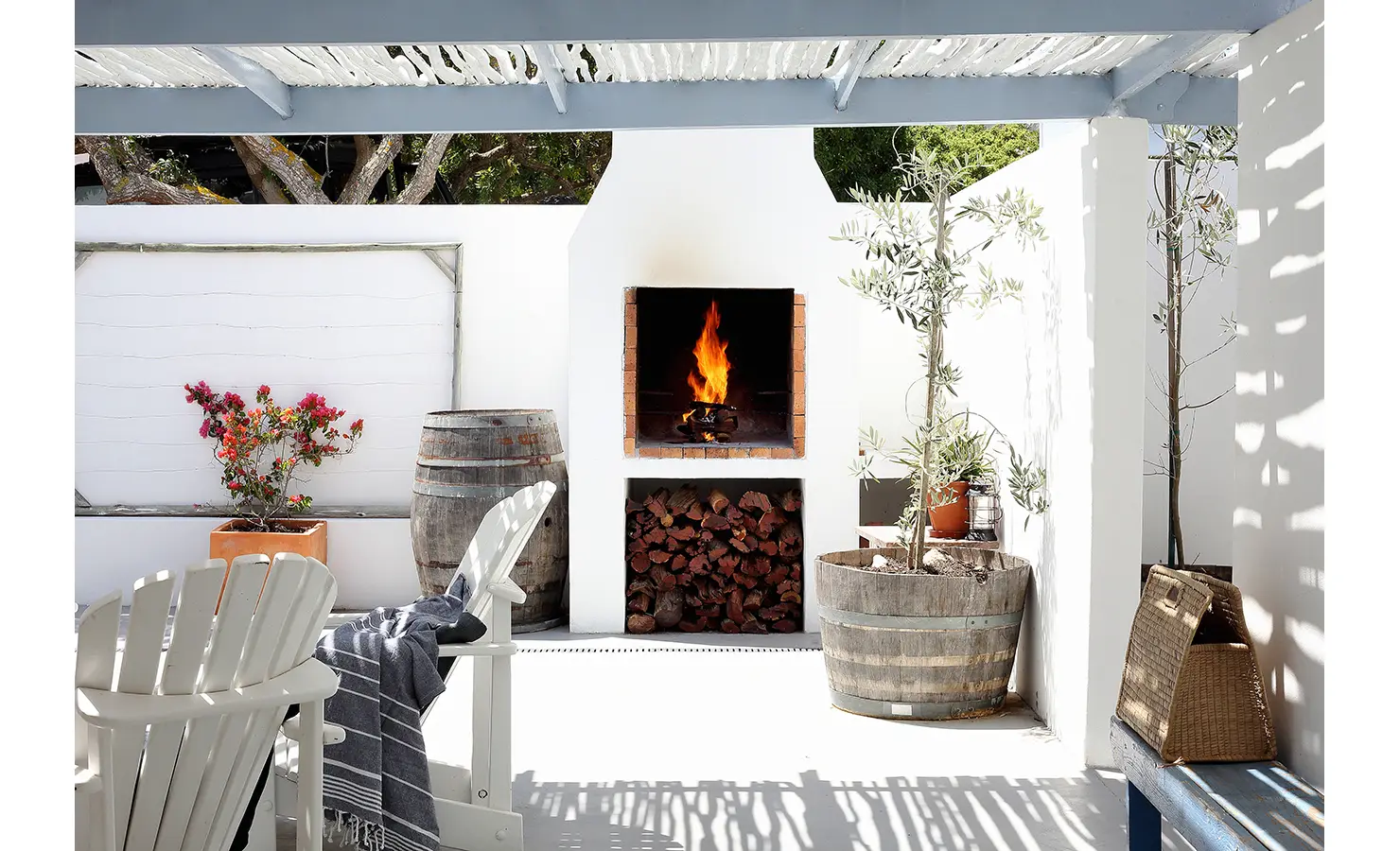 Perfect Hideaways: courtyard braai with roaring fire, stacked logs, olive trees in barrel planters and Adirondack chairs beneath a slatted pergola, Langebaan, Lagoon Lookout.