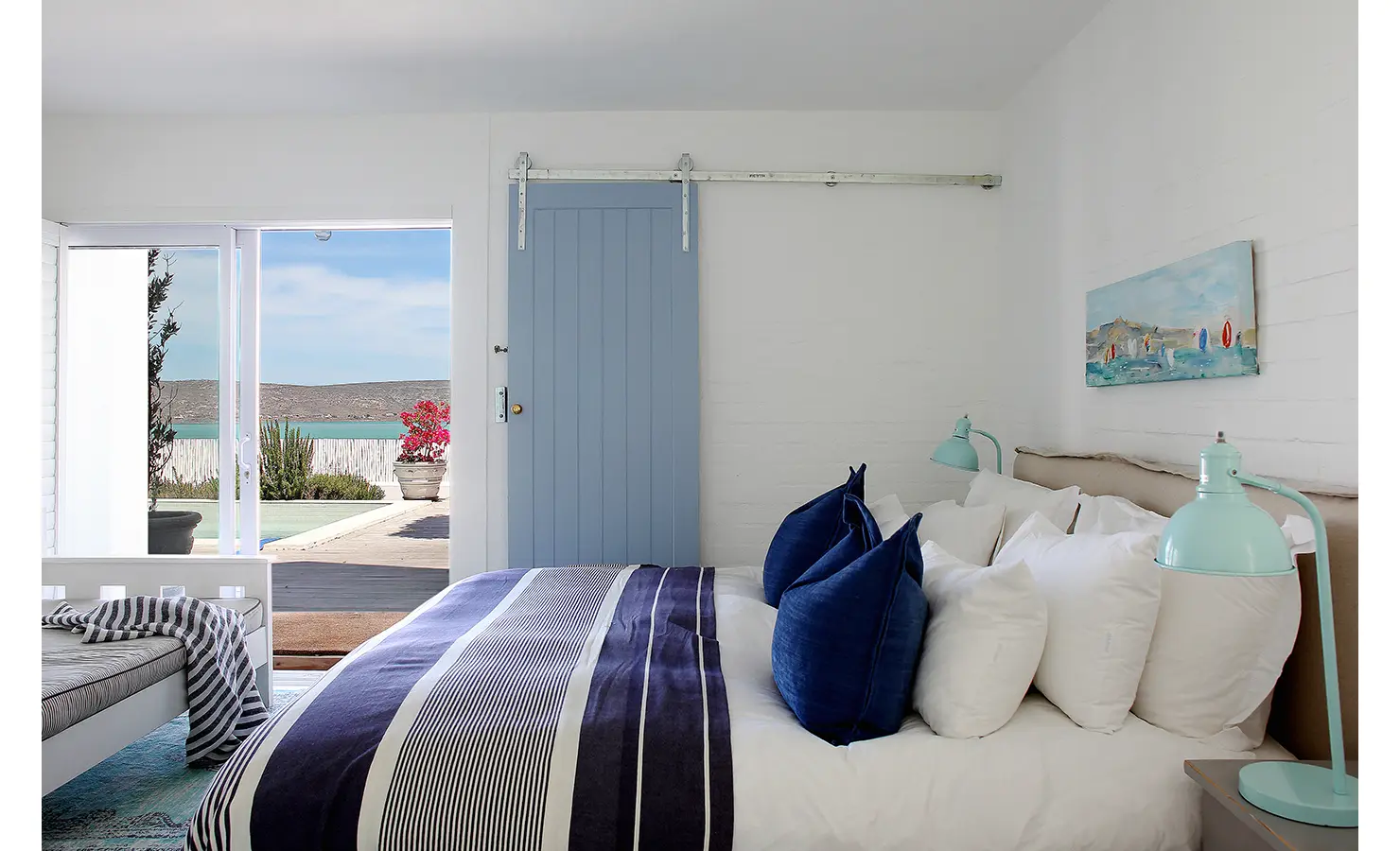 Perfect Hideaways: sea-facing bedroom with blue-striped bed, deep-blue cushions, mint lamps and a pale-blue barn door; doors open to the pool terrace and lagoon views, Langebaan, Lagoon Lookout.