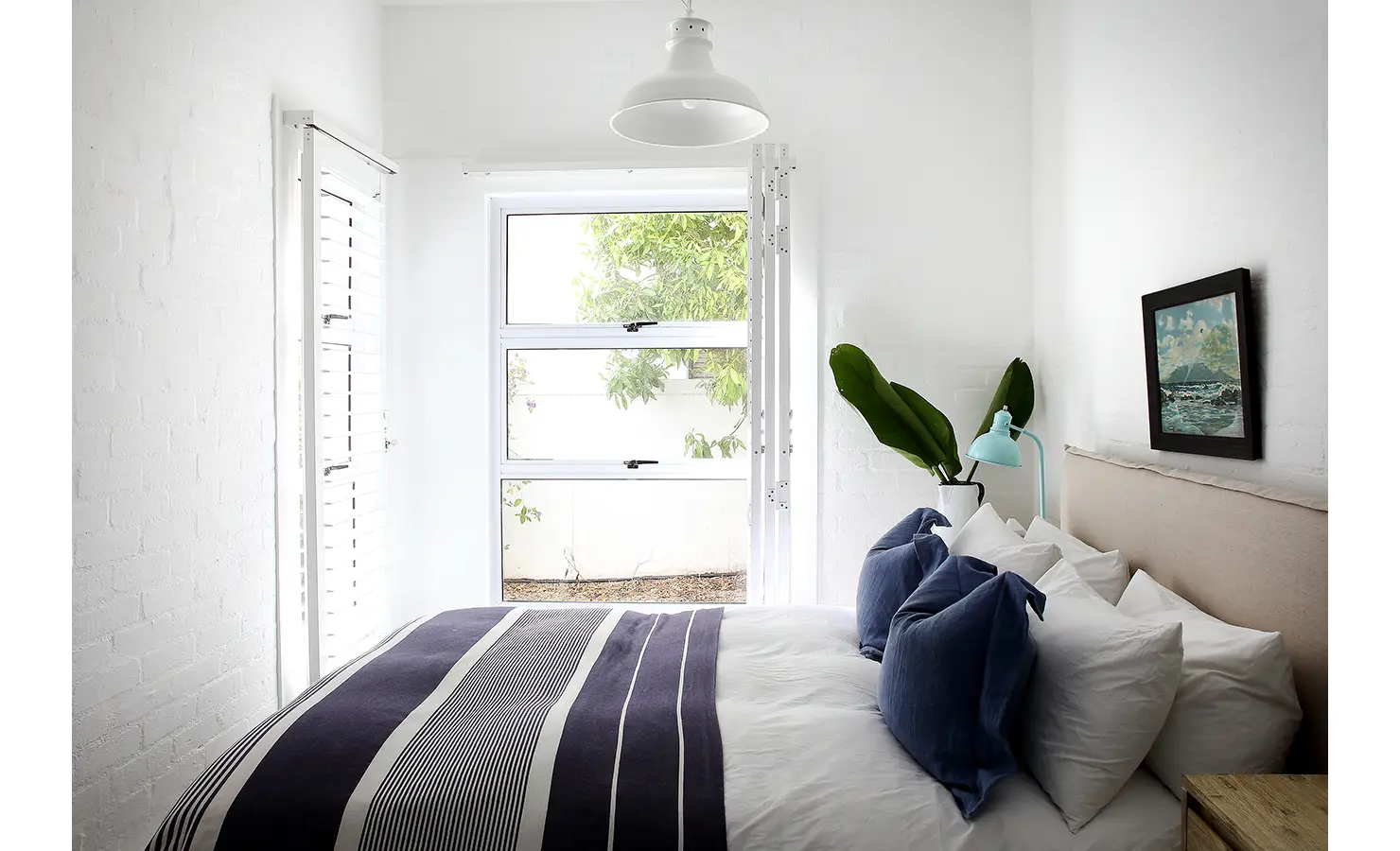 Perfect Hideaways: bright guest bedroom with a navy striped duvet, layered white pillows, coastal artwork and a shuttered window to courtyard greenery, Langebaan, Lagoon Lookout.