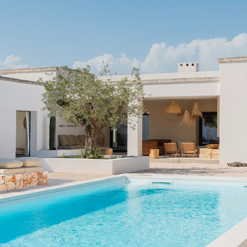 Perfect Hideaways whitewashed modern villa with a serene outdoor space featuring a turquoise swimming pool, an olive tree at the center of a sunlit courtyard, natural stone accents, and a shaded terrace with wooden furniture and woven pendant lights, evoking Mediterranean simplicity and calm. Ostuni, Casa Carbina.