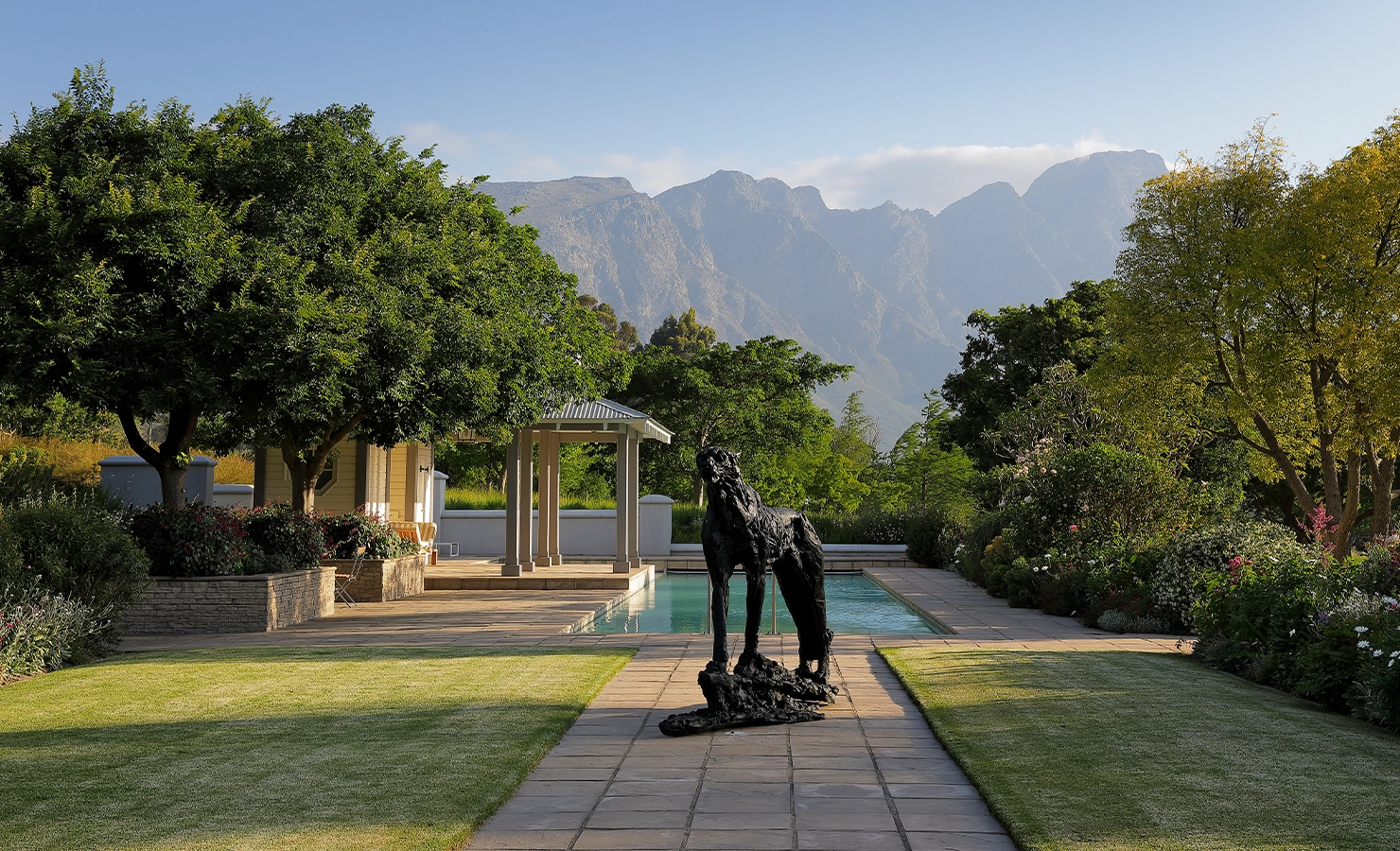 Perfect Hideaways serene garden scene featuring a long pool framed by manicured lawns, lush trees, and blooming shrubs, with a striking black sculpture of a dog in the foreground and dramatic mountain peaks rising in the distance under a clear blue sky, Franschhoek, Varuna.