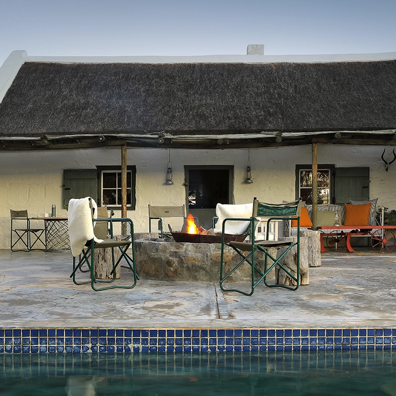 Perfect Hideaways - A rustic outdoor riverside picnic setup beside a classic open-top safari vehicle at Tankwa River Lodge. A red checked tablecloth covers a small table set with wine, drinks, and bowls, flanked by two green canvas chairs on stone slabs at the river’s edge. The calm water reflects the warm late-afternoon light, with golden bushes and distant mountains completing the tranquil Karoo landscape – Tankwa River Lodge, Tankwa Karoo.