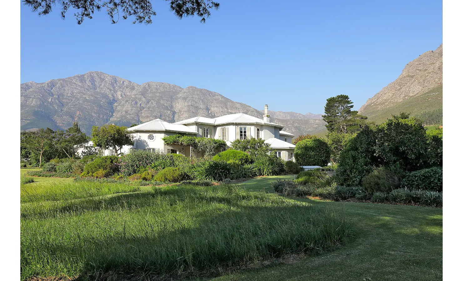 Perfect Hideaways elegant white manor nestled among lush gardens at the foot of rugged mountain peaks under a bright blue sky, Franschhoek, Varuna.