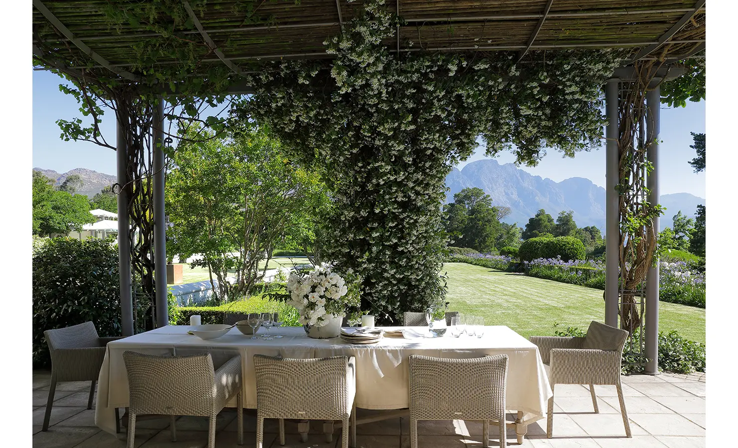 Perfect Hideaways shaded outdoor dining area with a table set beneath a pergola covered in flowering vines overlooking manicured lawns and mountains, Franschhoek, Varuna.
