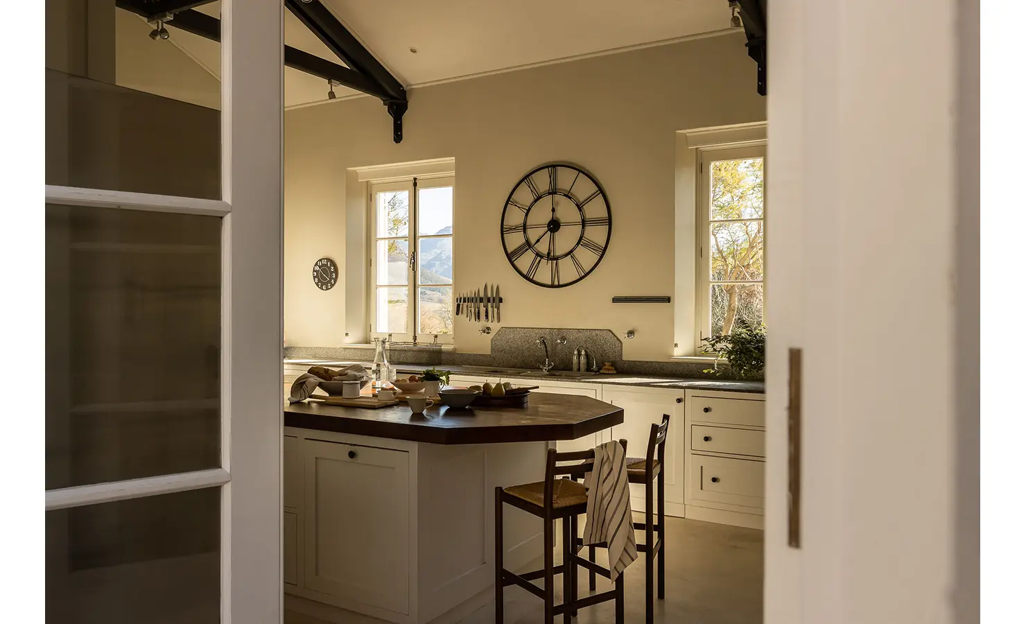 Perfect Hideaways warm, inviting kitchen with a central island, rustic bar stools, large clock on the wall, and sunlight streaming through tall windows, Franschhoek, Varuna.