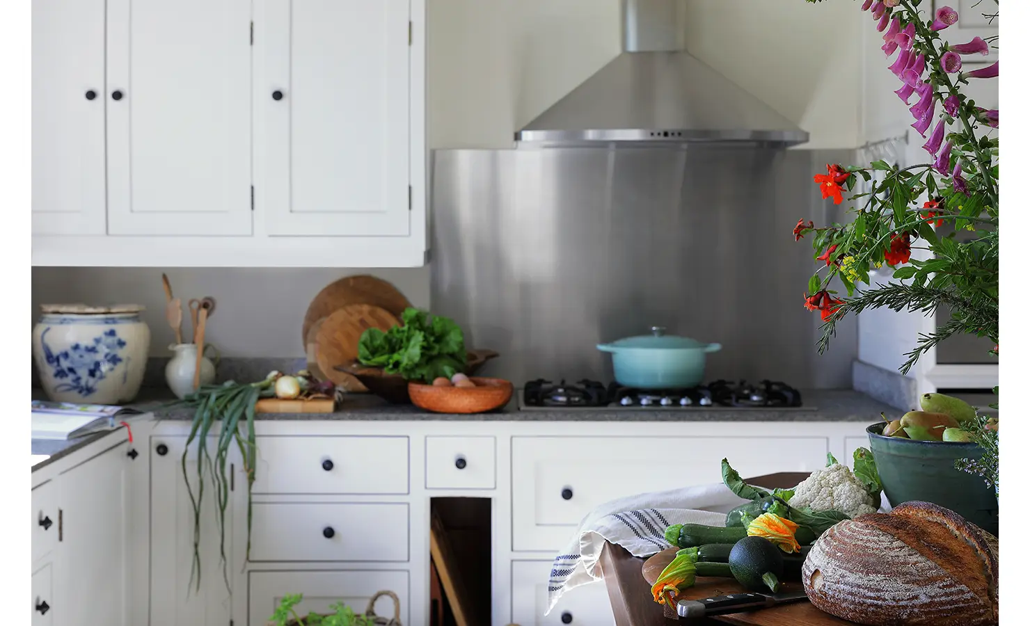 Perfect Hideaways bright farmhouse kitchen with white cabinetry, fresh vegetables, and bread on the counter beside colorful flowers and a blue cooking pot, Franschhoek, Varuna.