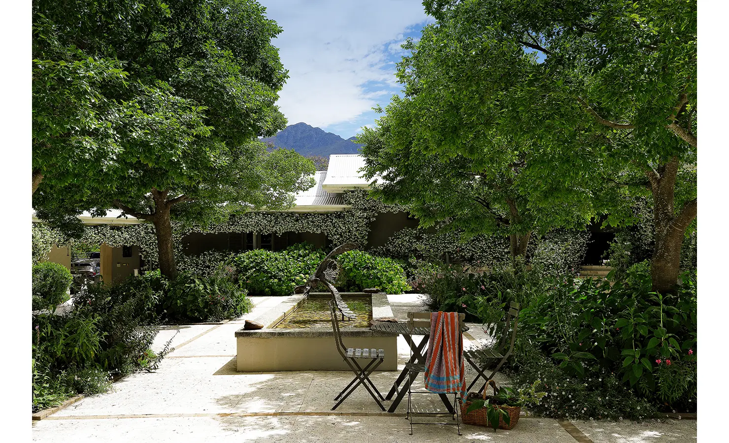 Perfect Hideaways shaded garden courtyard featuring a stone fountain, leafy greenery, and mountain views beyond the trees, Franschhoek, Varuna.