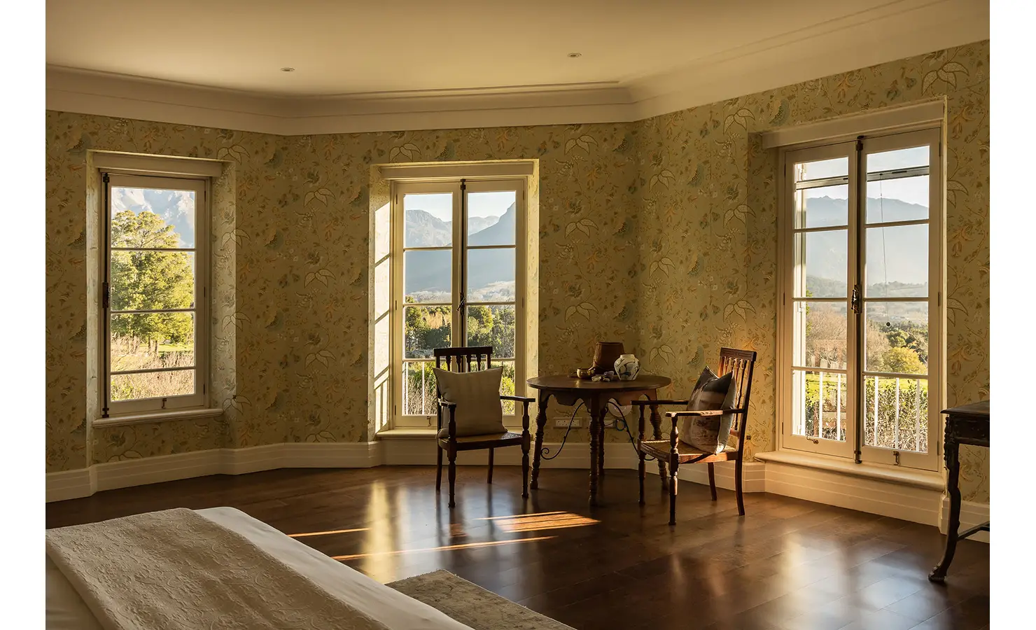 Perfect Hideaways serene bedroom with patterned wallpaper, large windows framing mountain scenery, and antique wooden chairs by a small table, Franschhoek, Varuna.